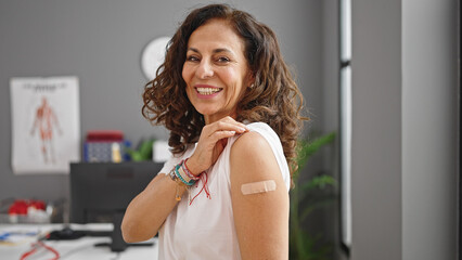 Middle age hispanic woman smiling confident standing with band aid on arm at clinic