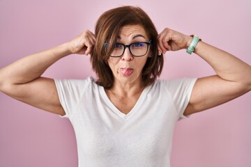 Middle age hispanic woman standing over pink background smiling pulling ears with fingers, funny gesture. audition problem