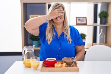 Obraz na płótnie Canvas Caucasian plus size woman eating breakfast at home smiling and laughing with hand on face covering eyes for surprise. blind concept.
