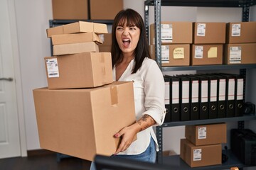 Young brunette woman working at small business ecommerce holding packages angry and mad screaming frustrated and furious, shouting with anger. rage and aggressive concept.