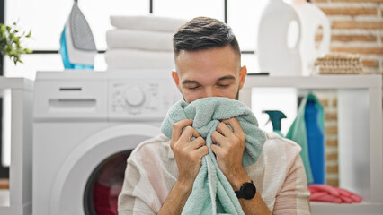 Young hispanic man smelling clean towel smiling at laundry room