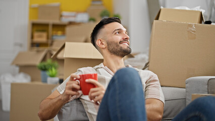 Young hispanic man drinking coffee sitting on floor at new home