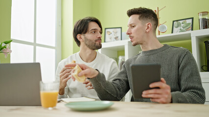 Two men couple having breakfast using touchpad and laptop at dinning room