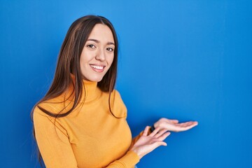 Fototapeta premium Young brunette woman standing over blue background inviting to enter smiling natural with open hand