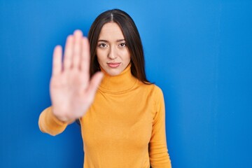Fototapeta premium Young brunette woman standing over blue background doing stop sing with palm of the hand. warning expression with negative and serious gesture on the face.
