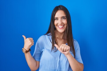 Fototapeta premium Young brunette woman standing over blue background pointing to the back behind with hand and thumbs up, smiling confident