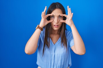Young brunette woman standing over blue background trying to open eyes with fingers, sleepy and tired for morning fatigue