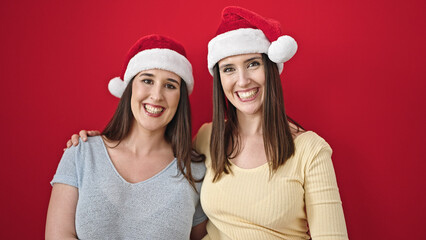 Two women hugging each other wearing christmas hat over isolated red background