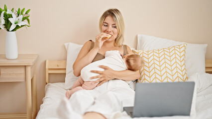 Mother and daughter breastfeeding baby watching movie on laptop eating croissant at bedroom