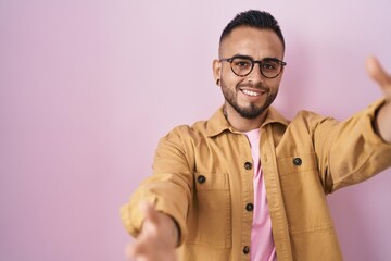 Young hispanic man standing over pink background looking at the camera smiling with open arms for hug. cheerful expression embracing happiness.