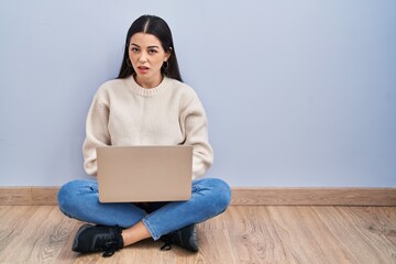 Young woman using laptop sitting on the floor at home in shock face, looking skeptical and...