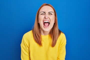 Young woman standing over blue background angry and mad screaming frustrated and furious, shouting with anger. rage and aggressive concept.