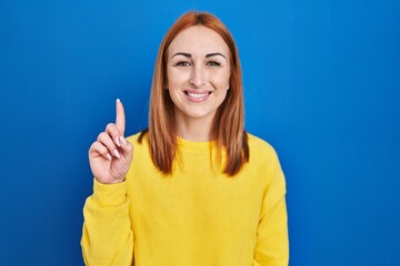 Young woman standing over blue background showing and pointing up with finger number one while smiling confident and happy.