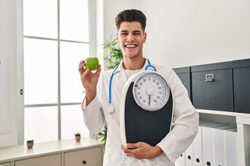 Young hispanic doctor man holding scale at dietitian clinic winking looking at the camera with sexy...