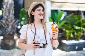 Young hispanic woman tourist holding camera eating ice cream at park