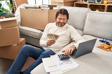 Middle age man using laptop counting denmark kroner banknotes at new home - obrazy, fototapety, plakaty