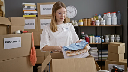 Young blonde woman volunteer packing clothes on cardboard smiling at charity center