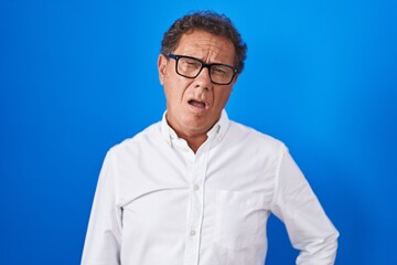 Middle age hispanic man standing over blue background in shock face, looking skeptical and sarcastic, surprised with open mouth
