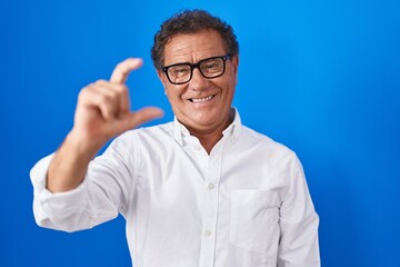 Fototapeta premium Middle age hispanic man standing over blue background smiling and confident gesturing with hand doing small size sign with fingers looking and the camera. measure concept.