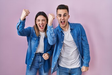 Young hispanic couple standing over pink background angry and mad raising fist frustrated and furious while shouting with anger. rage and aggressive concept.