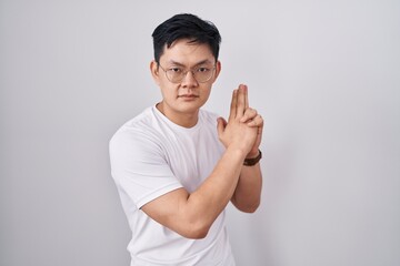 Fototapeta premium Young asian man standing over white background holding symbolic gun with hand gesture, playing killing shooting weapons, angry face