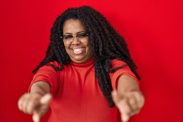 Plus size hispanic woman standing over red background pointing to you and the camera with fingers, smiling positive and cheerful