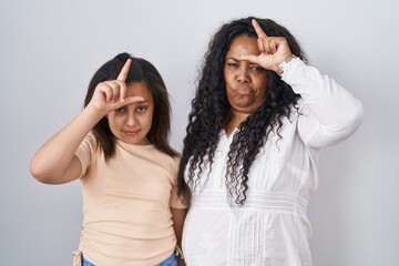 Mother and young daughter standing over white background making fun of people with fingers on forehead doing loser gesture mocking and insulting.
