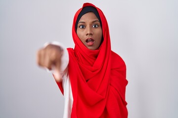 Young arab woman wearing traditional islamic hijab scarf pointing with finger surprised ahead, open mouth amazed expression, something on the front