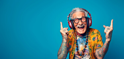 A happy hippie and cool grandfather, original style and tattoos, wearing headphones enjoying music, pointing his fingers up. Active and fun lifestyle concept for seniors: Sunset of life in colors - obrazy, fototapety, plakaty
