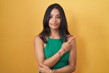 Obraz premium Brunette woman standing over yellow background pointing with hand finger to the side showing advertisement, serious and calm face