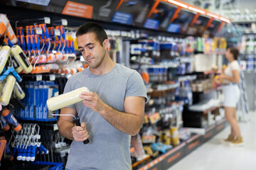 Focused interested young adult man standing next to rack stand with painting tools in construction...