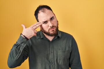 Plus size hispanic man with beard standing over yellow background shooting and killing oneself pointing hand and fingers to head like gun, suicide gesture.