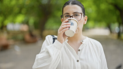 Young beautiful hispanic woman drinking take away coffee at the park