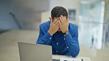 Young hispanic man business worker using smartphone and laptop stressed at the office