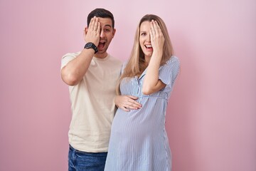 Young couple expecting a baby standing over pink background covering one eye with hand, confident...