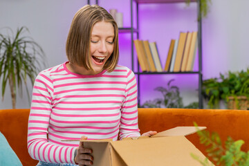 Happy brunette woman unpacking delivery parcel sits at home apartment on sofa. Smiling satisfied girl shopper online shop customer opening cardboard box receiving purchase gift by fast postal shipping