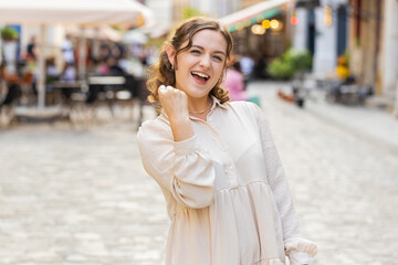 Fototapeta premium Portrait of young woman shouting, celebrating success, winning, goal achievement, good news, lottery jackpot luck, victory outdoors. Lovely pretty girl walking in urban city street. Town lifestyles