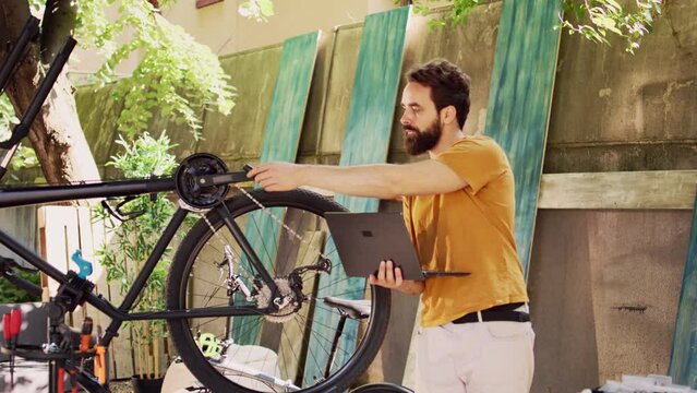 Sports-loving healthy man using laptop for enhancing safety of bike components for leisure cycling. Active male cyclist holding minicomputer for instructions to help repair damaged bicycle wheel.