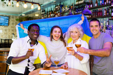 Happy man and woman sport fans holding flag of Ukraine, celebrating and drinking alcoholic drinks in beer pub, man showing victory sign