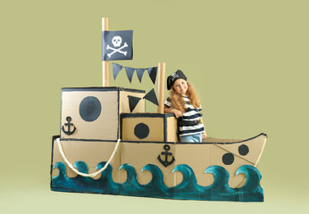 Cute little girl dressed as pirate in cardboard ship on green background