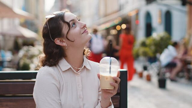 Happy young woman enjoying morning cold coffee drink with ice and smiling outdoors. Relaxing, taking a break. Girl sits on bench on city sunny street, drinking coffee to go. Town lifestyles outside