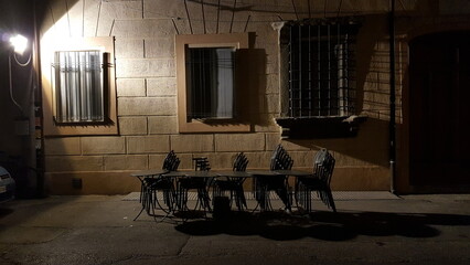 A wooden table and four chairs sit silently outside the building's entrance, illuminated only by the dim light of a nearby streetlamp..