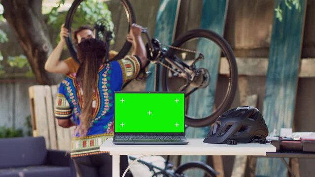 Laptop showing greenscreen while healthy and active interracial pair fixes bicycle components. Minicomputer with isolated copyspace screen on table while couple does bike maintenance at home yard.