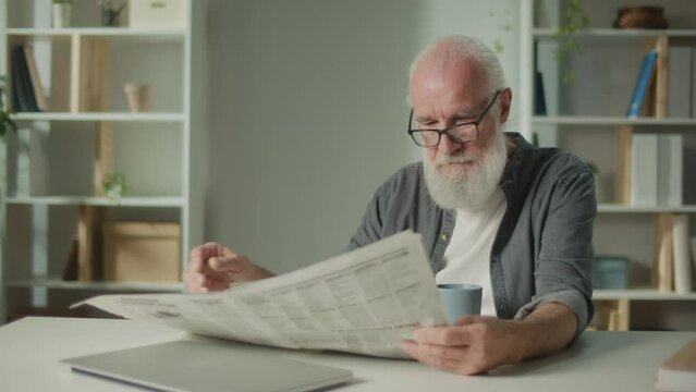 A Serious, Smart Old Man Sits at a Table and Reads a Newspaper.An Elderly Man with a Newspaper, Drinking Coffee Alone at Home, Sees the News and Current Events in the Newspaper.Daily Newspaper Concept