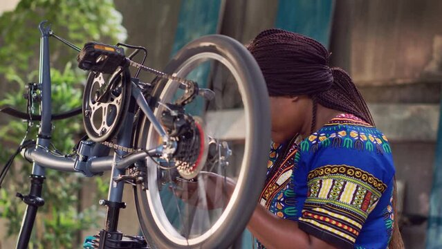 Outside close-up shot of young african american female securing and tightening bike wheel with professional equipment. Black woman holding expert tool for repairing modern bicycle outdoors.