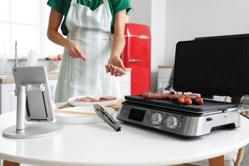 Young woman cooking sausages on electric grill in kitchen