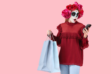 Shocked young woman with shopping bags and payment terminal on pink background. Mexico's Day of the Dead (El Dia de Muertos) celebration