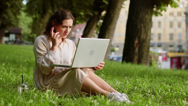 Portrait of young woman use laptop typing browsing working, loses becoming surprised sudden lottery results, bad news, fortune loss, fail. Girl sitting on grass in urban sunset city park background