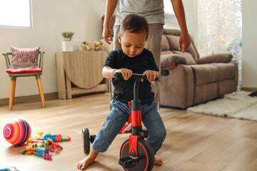 Adorable Asian little boy playing tricyle at home while his father watching and standing to protect him