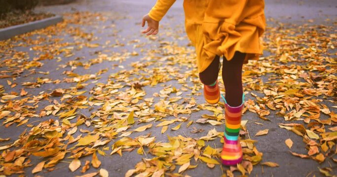 Autumn park and happy girl with colorful rubber boots and yellow dress running over fallen leaves on the ground